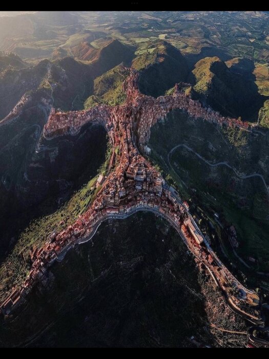 Historisk stad byggd längs en bergsrygg, omgiven av frodig natur och slingrande vägar. Ser ut som en flygbild.