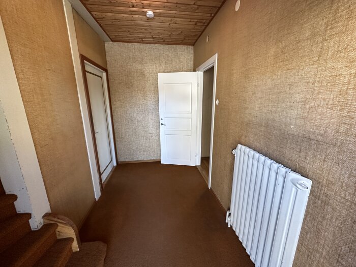 Trapphus med brunt golv, tapetserade väggar, vit dörr och radiator, trätrappsteg och paneltak.