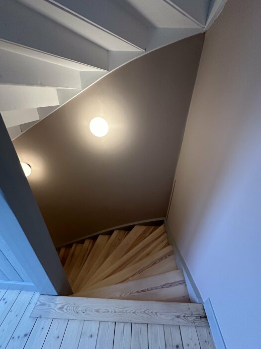 Trätrappa leder nedåt under sluttande tak med infällda spotlights och vita väggar.