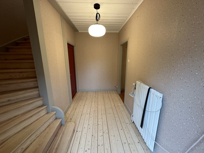 En tom hall med trätrappa, ljusa väggar, trägolv, radiator och taklampa.