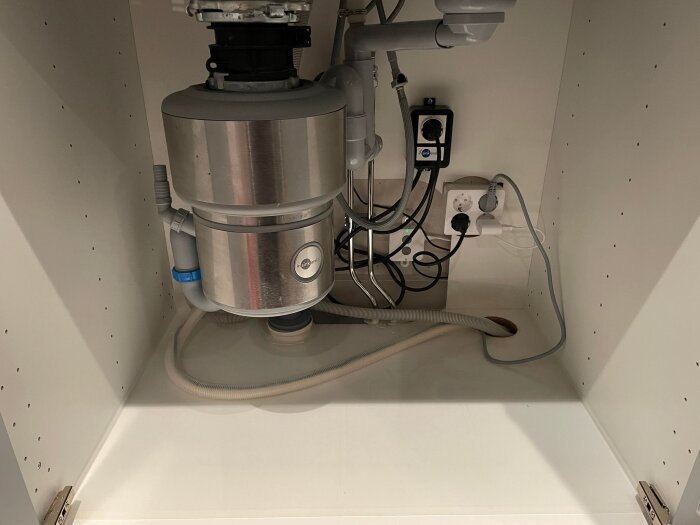 En avfallskvarn under diskbänk med sladdar och rörinstallationer, i ett skåp.