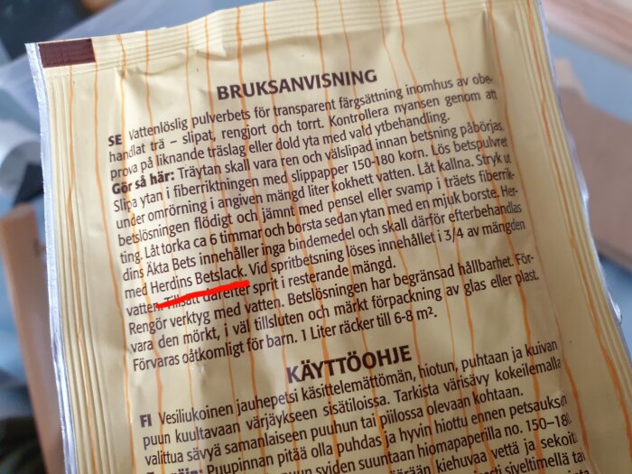 Bruksanvisning på behållare, möjligen färg eller bets, fokus på svensk text, något överstruket med rött.