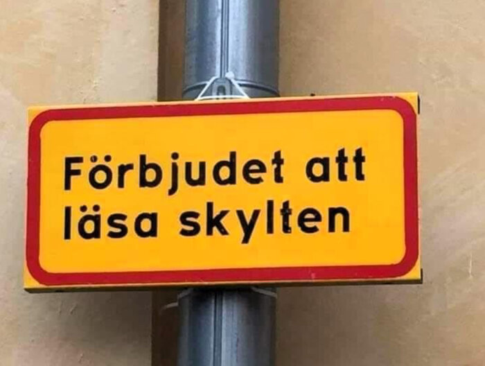 Gul skylt med text "Förbjudet att läsa skylten", paradoxalt budskap, fäst på stolpe.