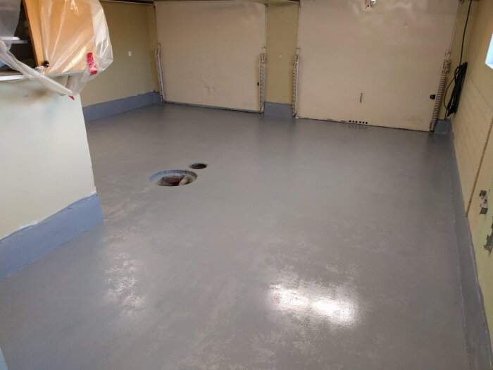 Ett tomt rum med grått golv, vita väggar och en öppen golvbrunn.