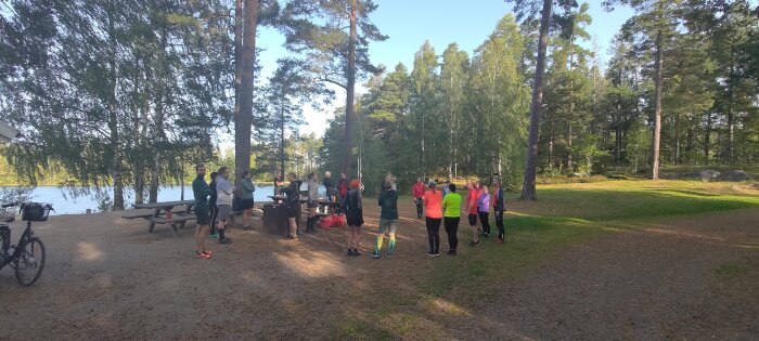 Grupp människor samlades i skogsparti nära vatten, soligt, diskuterar, idrottskläder, omgiven av natur och cykel.