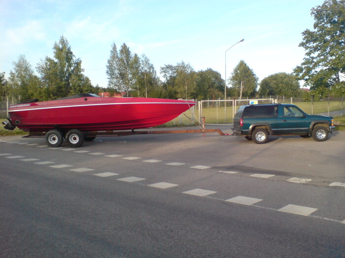 Röd båt på släpvagn, kopplad till SUV, parkerad vid vägkanten, dagtid, träd, lyktstolpe i bakgrunden.