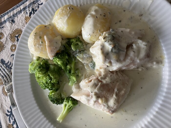 En tallrik med potatis, broccoli och kyckling i gräddsås. Hemlagad måltid på ett mönstrat underlägg.