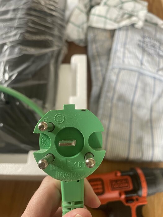 En hand håller en grön kontakt med skruvar och tråd, otydlig bakgrund med verktyg och textilier.