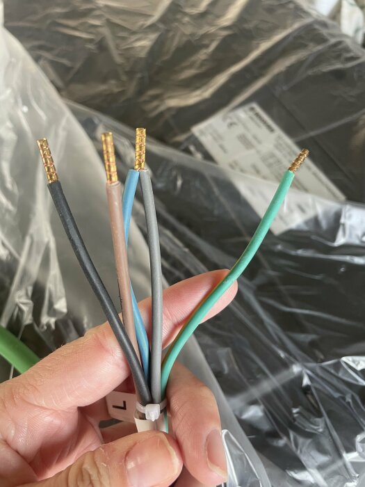 Hand håller fem avskalade elektriska kablar med olika färger framför en plastpåse.