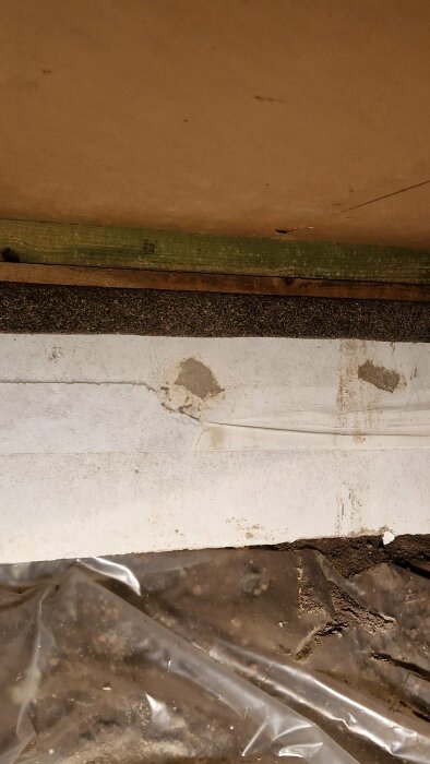 Bild av en hörnsektion med smutsiga ytor, spånskiva, vit vägg och plastfolie på golvet. Slitage synligt.