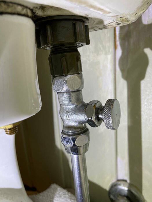 Toalettens vattenförsörjning med avstängningsventil och anslutningsrör under handfat, något kalk- och rostbelagd.