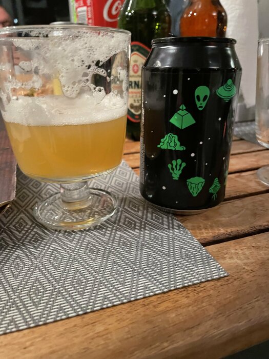Ett ölglas med skum, en burk med utomjordingstryck, flaskor i bakgrunden, på ett träbord.