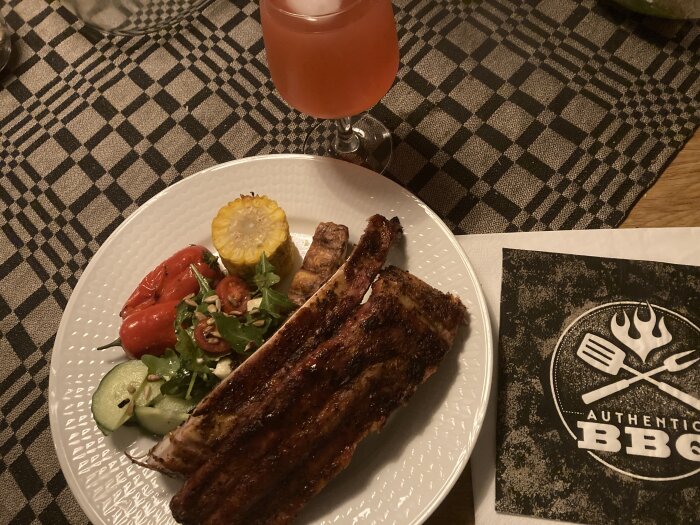 Grillad fläskkotlett, sallad, grillad citron, röd dryck och BBQ-restaurangservett på rutig duk.