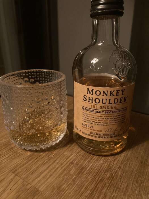 Tömt whiskyglas bredvid en öppnad flaska Monkey Shoulder skotsk whisky på ett bord.