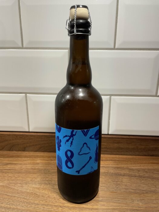 Brun glasflaska med flip-top kork och blå etikett mot kakelvägg och träyta.