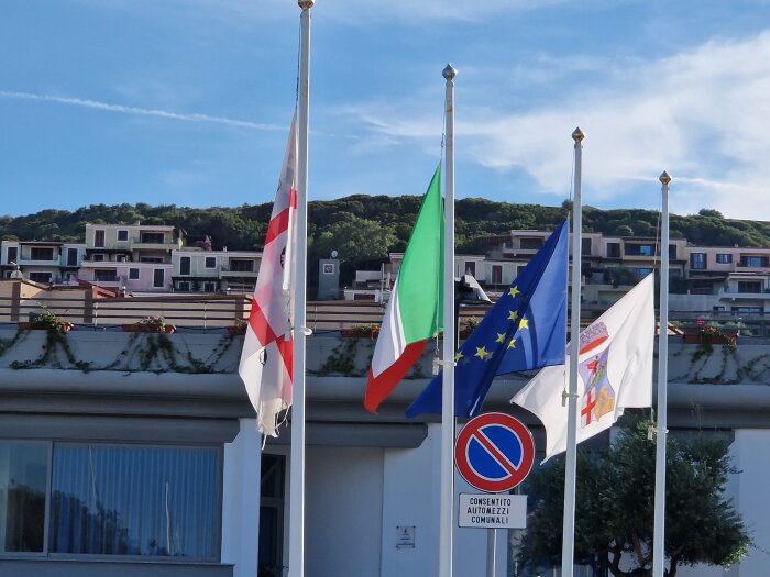 Flaggor framför byggnad, Italien och EU bland andra, klart väder, grönska, moderna bostadshus i bakgrunden.