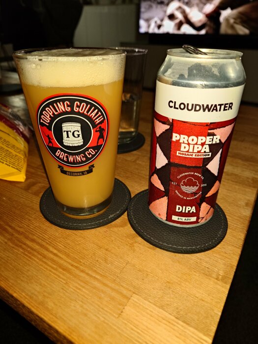 Ölglas på disk med logotyp, ölburk med etikett "Proper DIPA", träyta, suddig bakgrund med TV-skärm.
