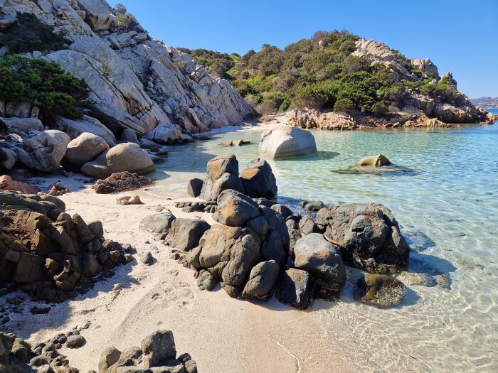 Klart turkost vatten, sandstrand, klippor, grönska, blå himmel, orörd natur, lugn, vacker kustmiljö.