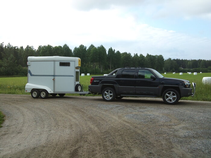 SUV kopplad till en hästtrailer parkerad på en grusväg med fält och skog i bakgrunden.