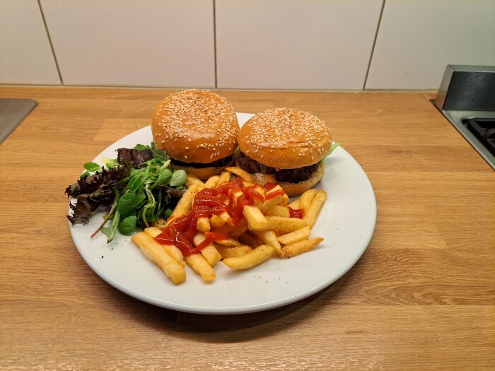 Två hamburgare med sesambröd, pommes frites, grönsallad och ketchup på en vit tallrik.