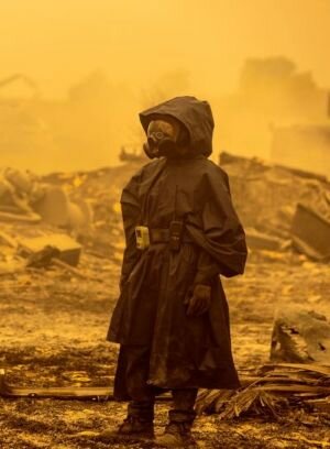 Person i skyddsdräkt och gasmask står i förödelse under gulaktig himmel, möjligen efter brand eller katastrof.