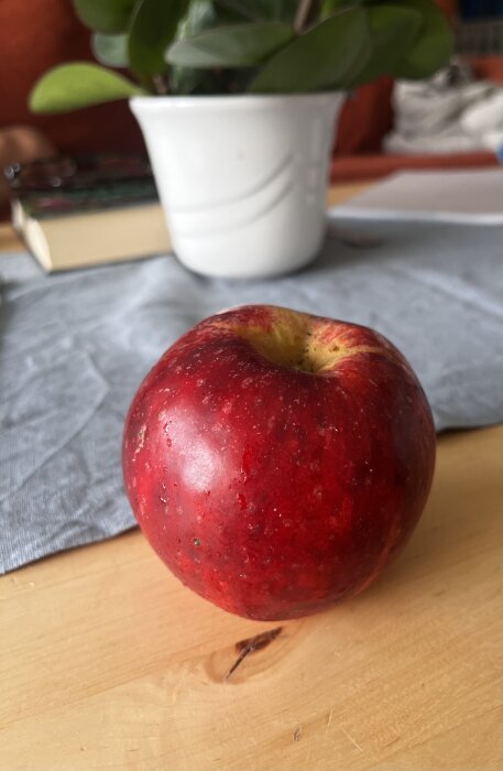 Rött äpple på träbord, otydlig växt och bok i bakgrunden, grått tyg under äpplet.