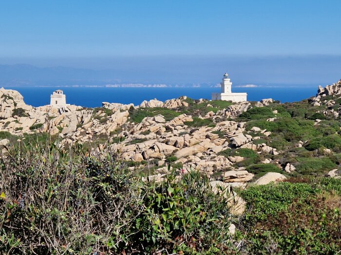 Kustlandskap med fyr, klippor, vegetation och ett klart blått himmel.