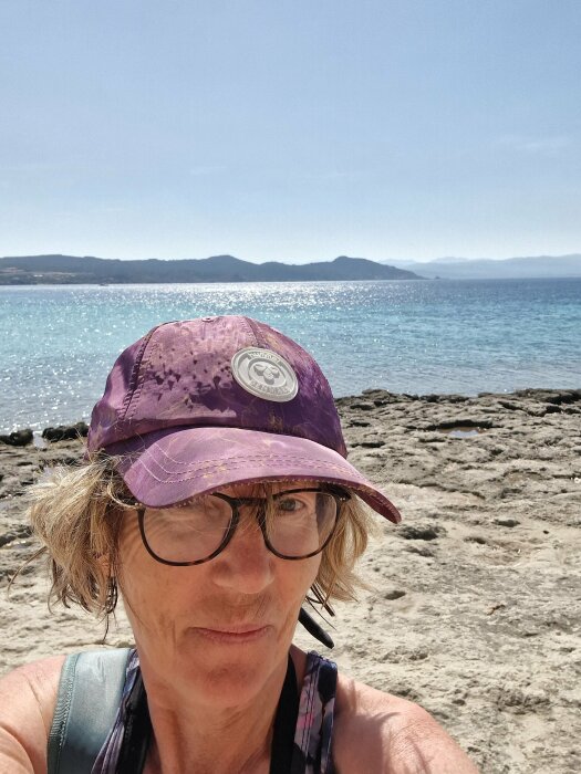 Person med keps framför havet, soligt väder, klippig strandkant, dagtid.