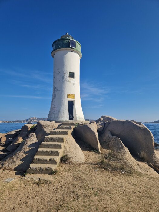 Vit fyr vid havet, klarblå himmel, soligt, stentrappor, stora klippblock, gräsmatta, inga personer, dagtid.