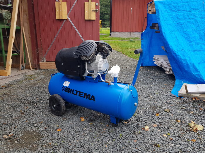 Blå kompressor från Biltema utomhus på grus, med byggnader och blå presenning i bakgrunden.