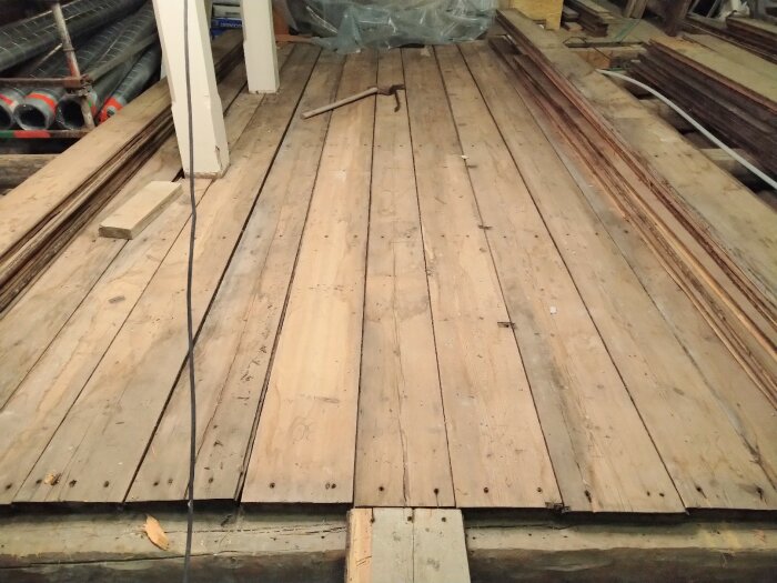 Trägolv i byggnad med byggmaterial, rör och konstruktionsstänger. Oordnat, pågående arbete eller renovering.