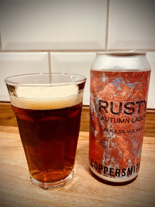 Ett glas med bärnstensfärgad dryck bredvid en ölburk. "Rusty Autumn Lager," träyta, inget folk.