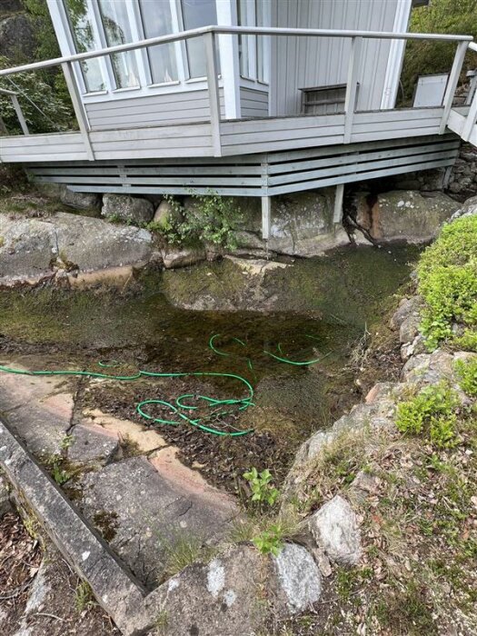 Trädäck på stolpar över klippig mark med grön vattenslang i en liten grop med vatten.