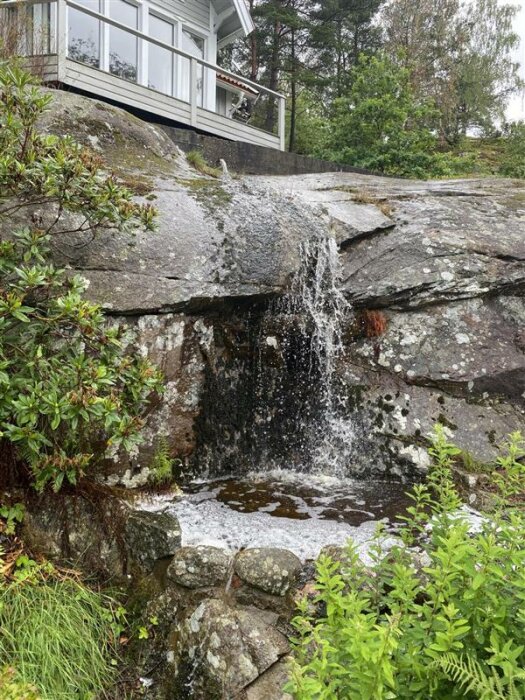 Ett litet vattenfall vid klippor under ett hus med veranda, omgivet av grönska i ett skogslandskap.