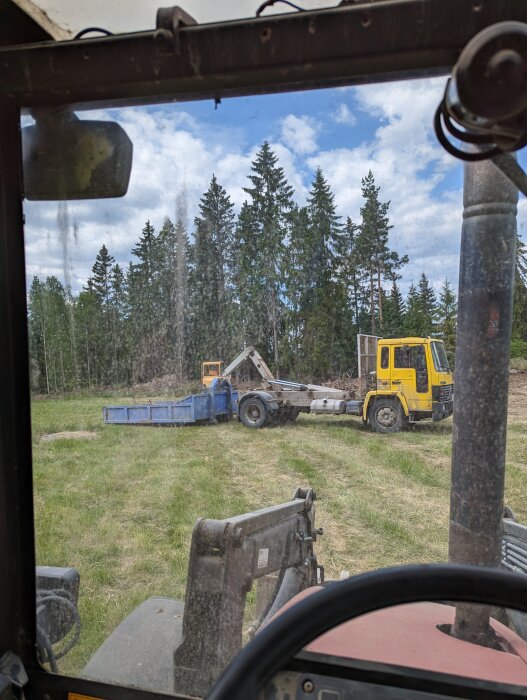 Utsikt från traktorns hytt: skogsbakgrund, smutsigt fönster, gul lastbil, blå släpvagn, gräsmark.