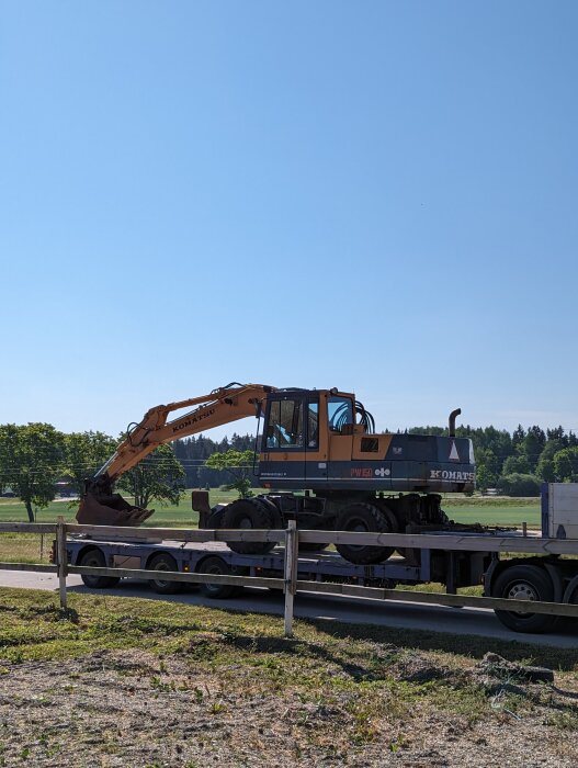 Grävmaskin på lastbilstrailer, parkerad vid väg, klart väder, ingen person synlig.