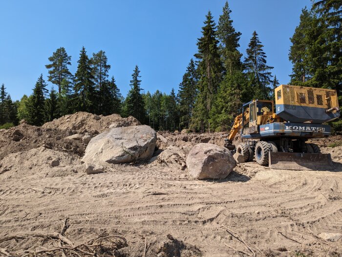 En byggarbetsplats med en Komatsu grävmaskin, stora stenblock, högar av jord och skog i bakgrunden.