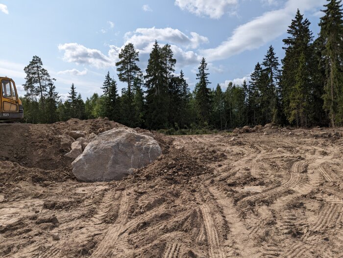 Markarbetsplats med grävmaskin, stor sten, skogsbakgrund och spår i sanden under solig himmel.