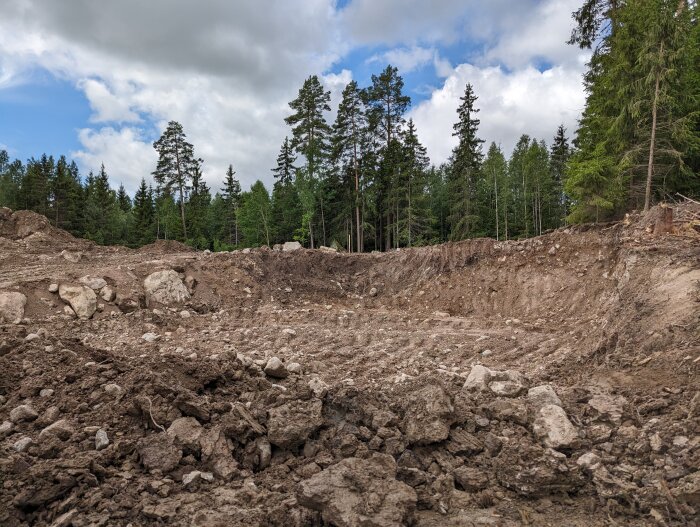 Uppgrävd mark, stenar, skogsbakgrund, molnig himmel, potentiell byggplats eller gruvaktivitet.