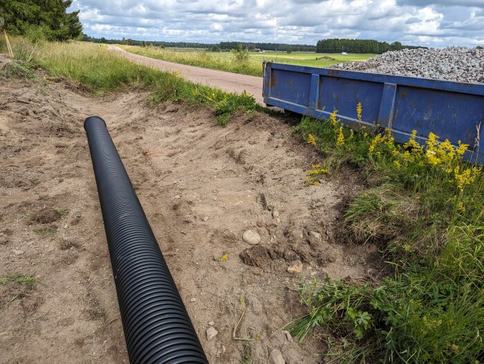 Lantväg, grävning, svart dräneringsrör, blå container med grus, gröna fält, delvis molnig himmel.