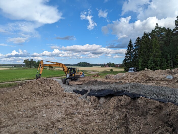 Grävmaskin vid jordhög, byggarbetsplats, gröna fält i bakgrunden, delvis molnig himmel.