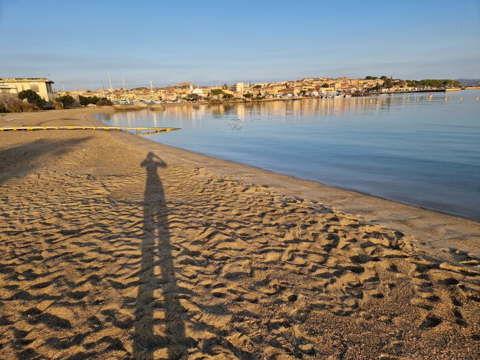 Strand, hav, sand, skugga av person, båtar, byggnader i bakgrunden, soligt, klart väder, lugnt vatten.