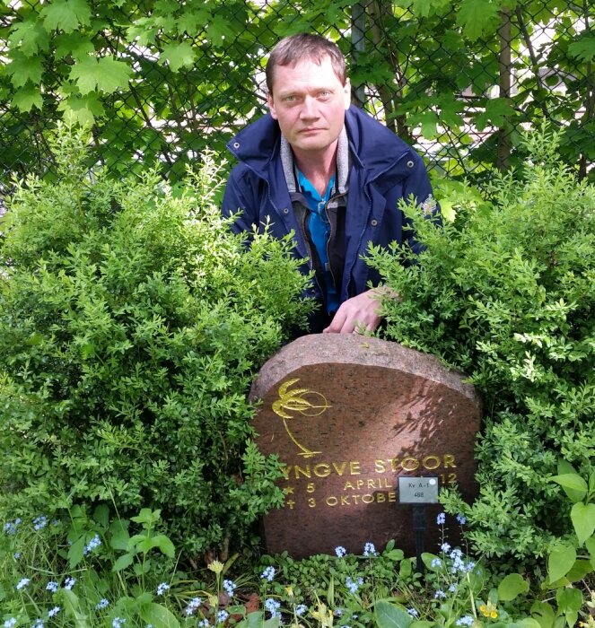 Man står bakom gravsten omgiven av gröna växter och blommor.