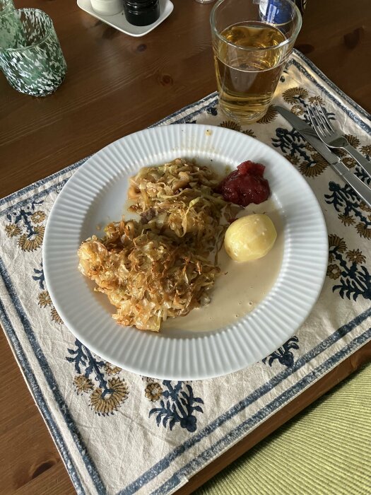 Måltid på bord: kålpudding, potatis, lingonsylt, sås, dricksglas med läsk, gaffel, duk, lantlig inredning.