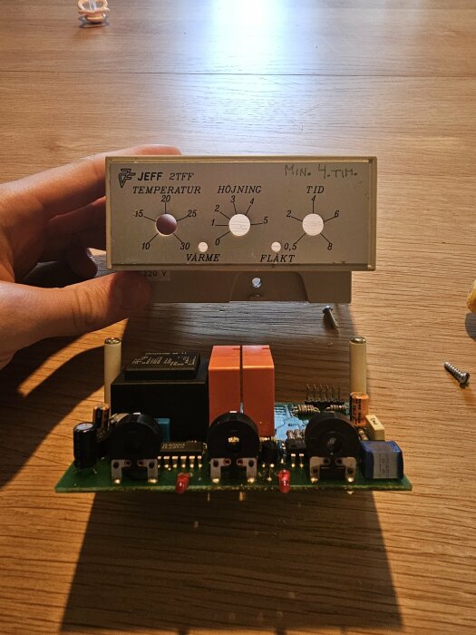 Elektronisk kretskort med reglage för temperatur och fläkthastighet, hand som håller, bord i bakgrunden.