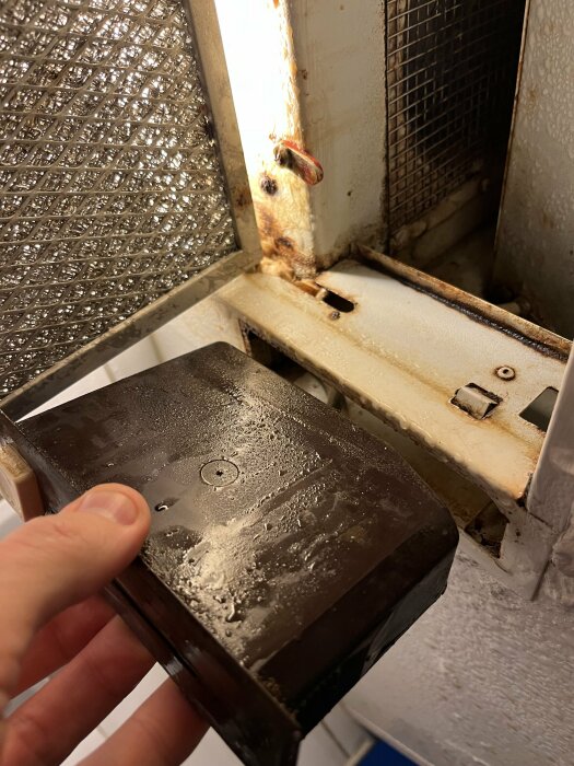 Hand håller en isig del framför en öppen frys, kondens och frost, nedslitet, behöver troligen rengöring eller underhåll.