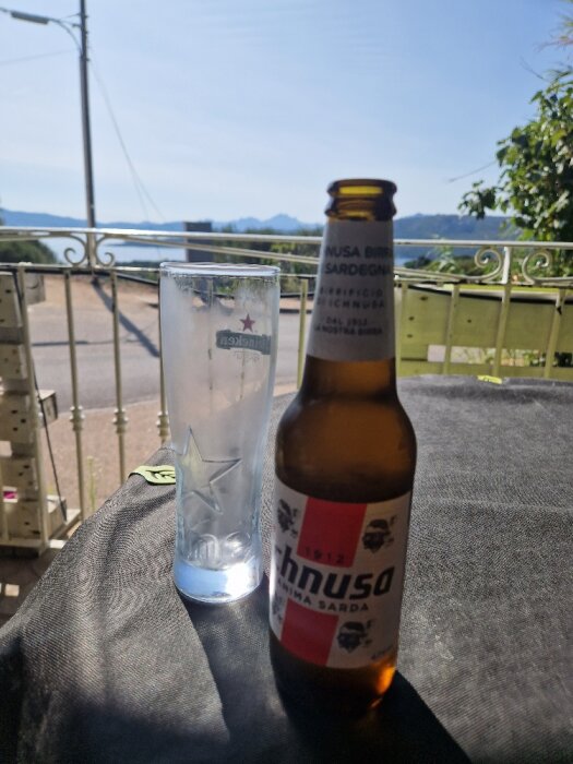 Ölflaska och tomt glas på bord framför vacker utsikt, dagtid, klar himmel, berg i bakgrunden.