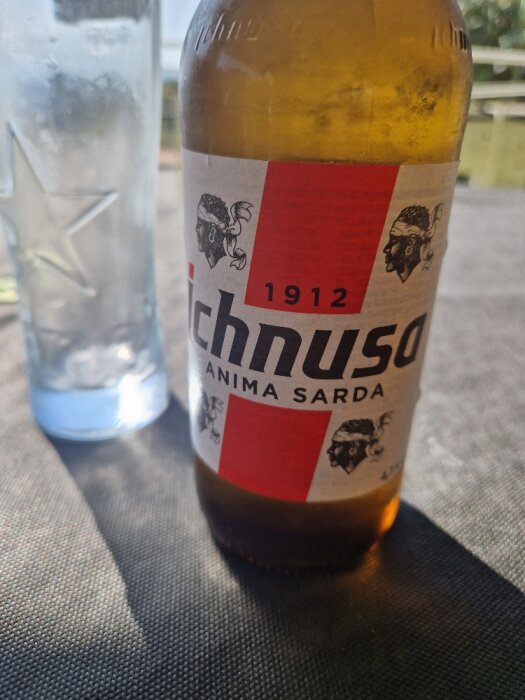 En ölflaska med etikett "Ichnusa" framför en suddig vattenflaska, på ett bord, utomhus i dagsljus.