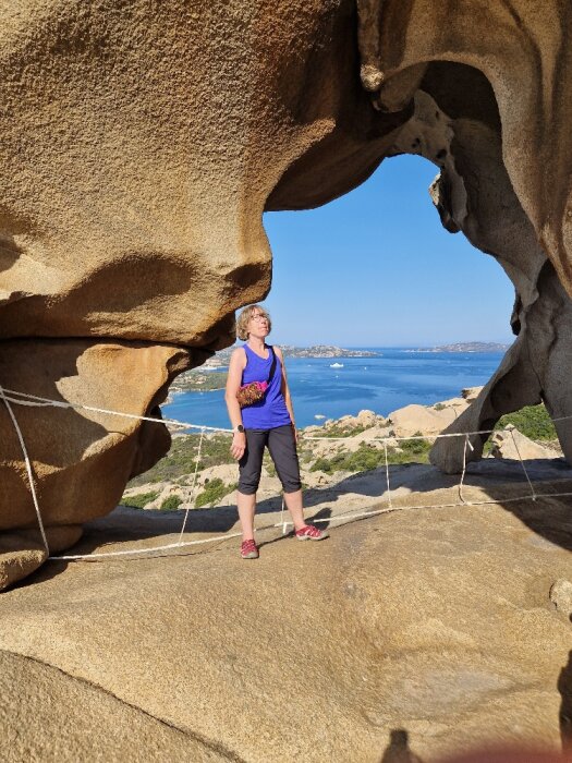 Kvinna står under naturlig stenformation, klar himmel, hav i bakgrunden, soligt, rep som skydd.