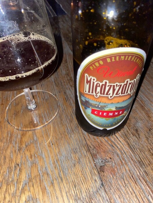 Flaska mörk öl med etikett "Międzyzdroje", halvfylld ölglas, träbord, fokus på framträdande etikett.